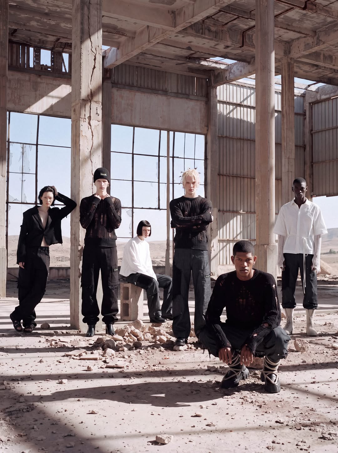 Six models in black and white garments standing among the pillars of an abandoned concrete building in the desert.
