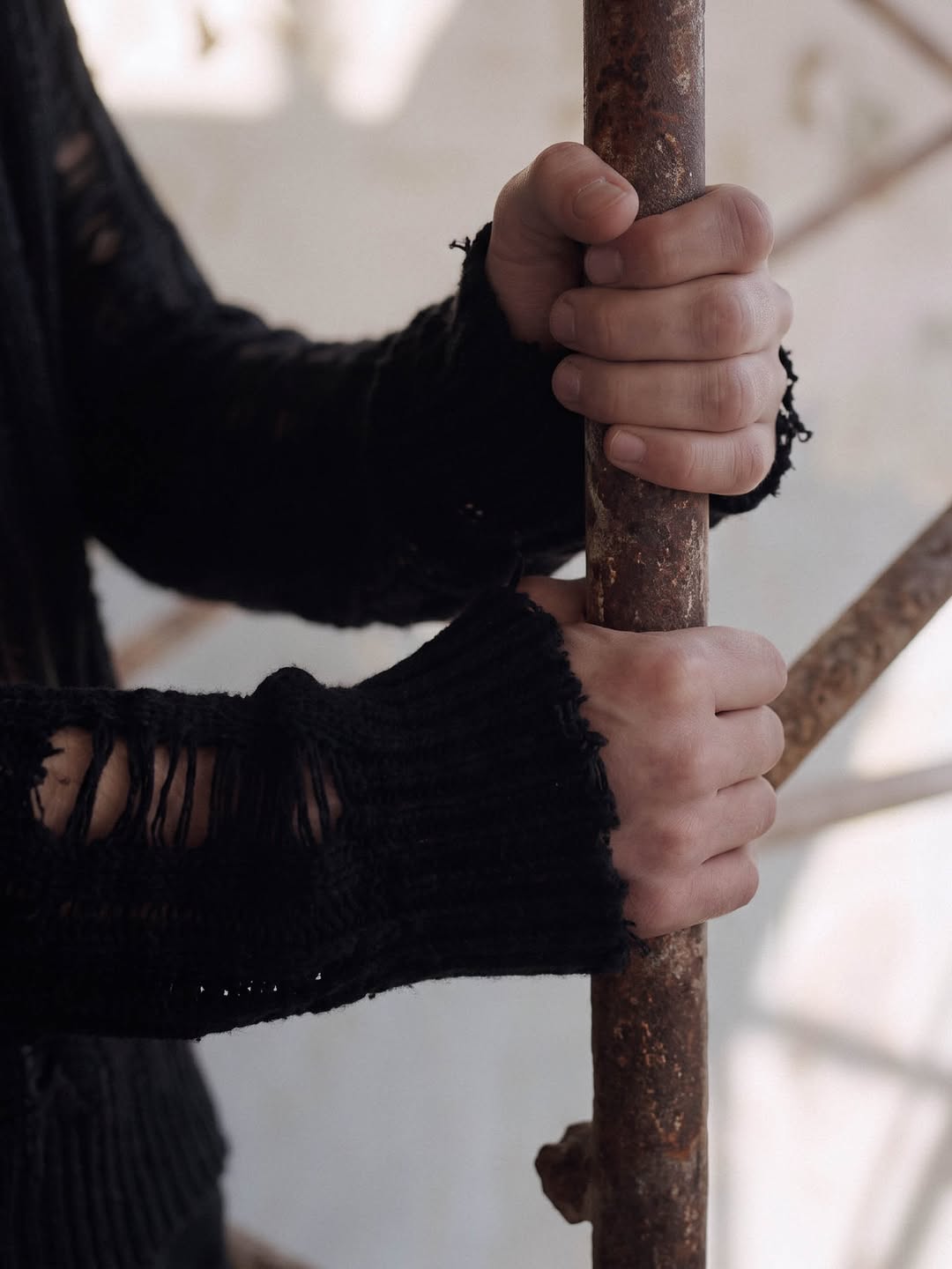 Hands in frayed black knit cuffs gripping a rusted iron pole.