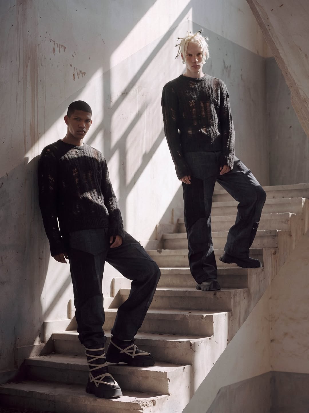Two models in distressed black knits on a concrete staircase, window light falling across the wall.