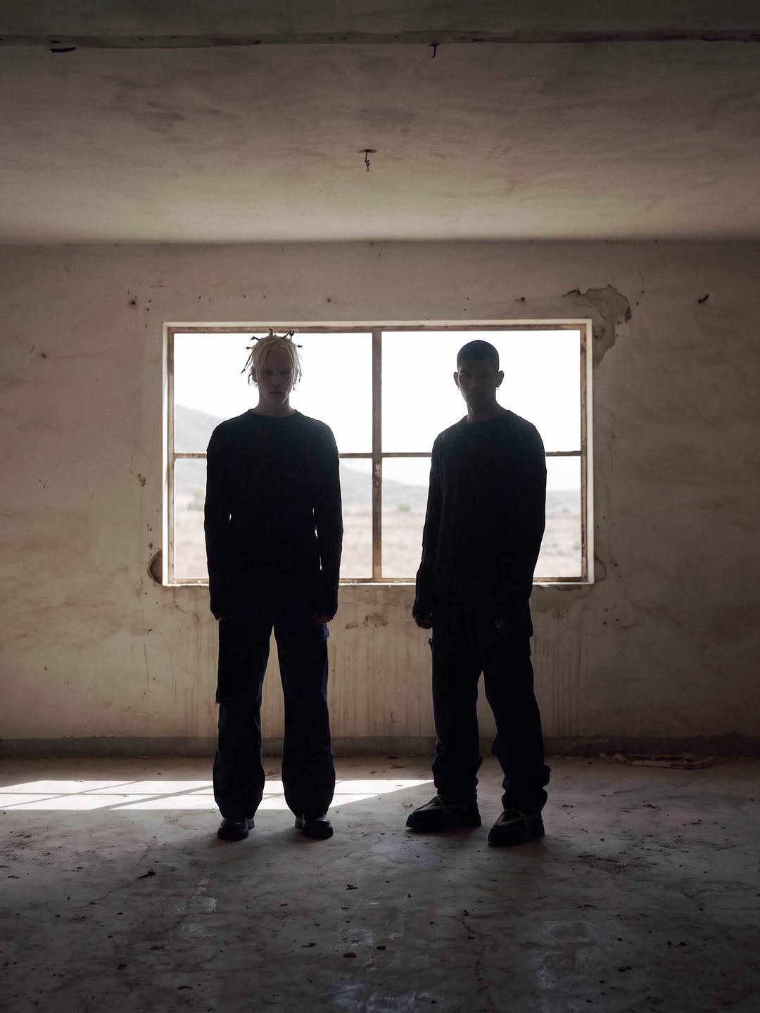 Two figures in black open-knit sweaters and wide denim, standing against a window in a bare concrete room.